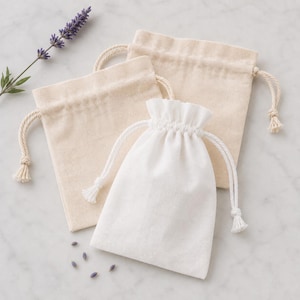 Può includere: Due sacchetti con coulisse beige e un sacchetto con coulisse bianco in tessuto. Ogni sacchetto presenta una chiusura con cordino intrecciato abbinato con estremità annodate. Sono visibili un rametto di lavanda viola e alcuni chicchi sparsi.