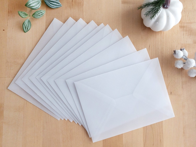 May include: A stack of white envelopes on a wooden surface. The envelopes are arranged in a fan shape, with the top envelope being the most visible. The envelopes are all the same size and shape, and they have a smooth, matte finish.