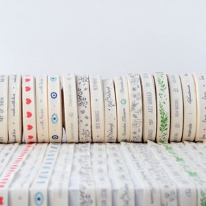 May include: A collection of cream-colored ribbons with various printed designs and text. Designs include hearts, floral patterns, and phrases like "Thank You" and "Just Married." Some ribbons have red, blue, and green accents.