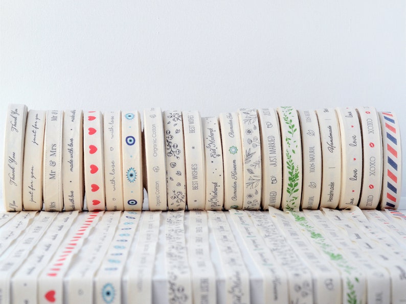 May include: A collection of cream-colored ribbons with various printed designs and text. Designs include hearts, floral patterns, and phrases like "Thank You" and "Just Married." Some ribbons have red, blue, and green accents.