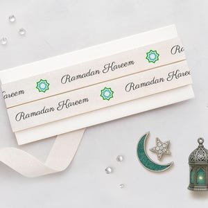 May include: Cream-colored ribbon with the text "Ramadan Kareem" and a green and blue starburst design. The ribbon is wrapped around a stack of white cards. A crescent moon and star pin and a decorative lantern are also present.