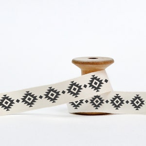 May include: A spool of white cotton ribbon with a black geometric pattern. The ribbon is wound around a wooden spool.