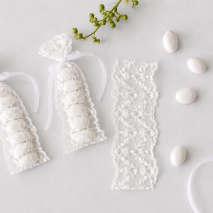 Op de afbeelding: Twee witte kanten zakjes gevuld met witte amandelsnoepjes, vastgebonden met dunne witte linten. Een strook bijpassend wit kant, vijf losse witte amandelsnoepjes, een enkel wit lint en een klein groen takje liggen erbij.