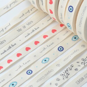 May include: Cream-colored ribbon rolls with various printed designs. The ribbons feature phrases like "Thank You," "Mr & Mrs," and "Best Wishes," along with red hearts, blue evil eye symbols, and floral patterns. The text "Organic Cotton" is also printed on the ribbon.