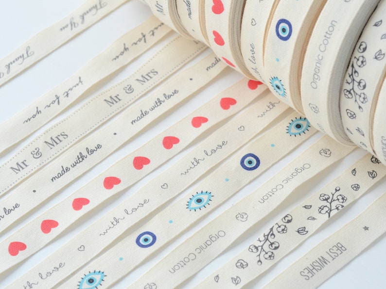 May include: Cream-colored ribbon rolls with various printed designs. The ribbons feature phrases like "Thank You," "Mr & Mrs," and "Best Wishes," along with red hearts, blue evil eye symbols, and floral patterns. The text "Organic Cotton" is also printed on the ribbon.