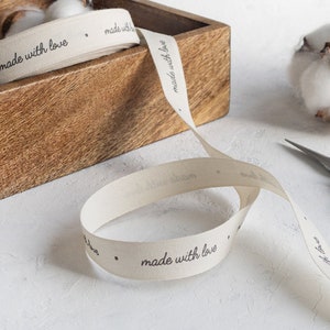 May include: Cream-colored ribbon with the words "made with love" printed in black. The ribbon is partially rolled in a wooden box. A pair of scissors and cotton buds are also visible.