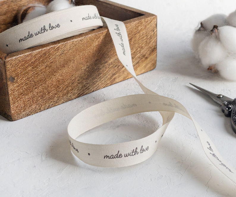 May include: Cream-colored ribbon with the words "made with love" printed in black. The ribbon is partially rolled in a wooden box. A pair of scissors and cotton buds are also visible.