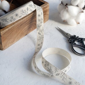 Puede incluir: Una cinta blanca con un patr&oacute;n floral negro est&aacute; envuelta alrededor de una caja de madera. La cinta tambi&eacute;n est&aacute; enrollada sobre una superficie blanca junto a un par de tijeras plateadas.