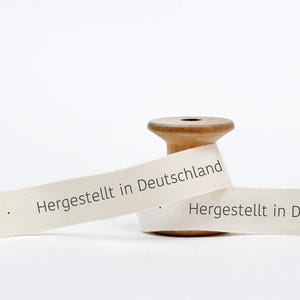 May include: A spool of white ribbon with the text "Hergestellt in Deutschland" printed on it. The ribbon is wrapped around a wooden spool.