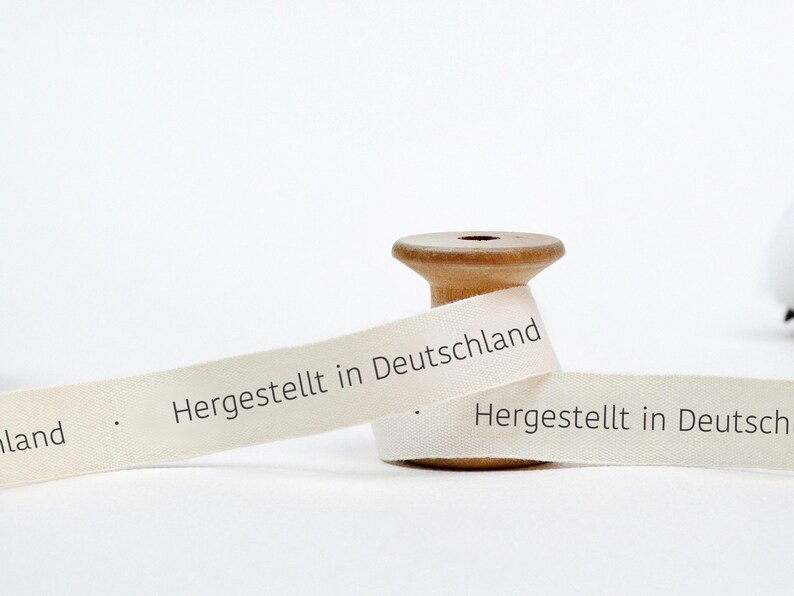 May include: A spool of white ribbon with the text "Hergestellt in Deutschland" printed on it. The ribbon is wrapped around a wooden spool.