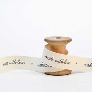 May include: A wooden spool with cream-colored ribbon. The ribbon is printed with the words "made with love" in black script. The spool is light brown and has a hole in the center.