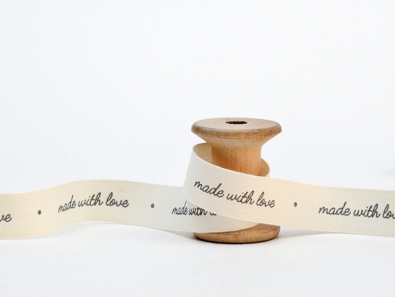May include: A wooden spool with cream-colored ribbon. The ribbon is printed with the words "made with love" in black script. The spool is light brown and has a hole in the center.