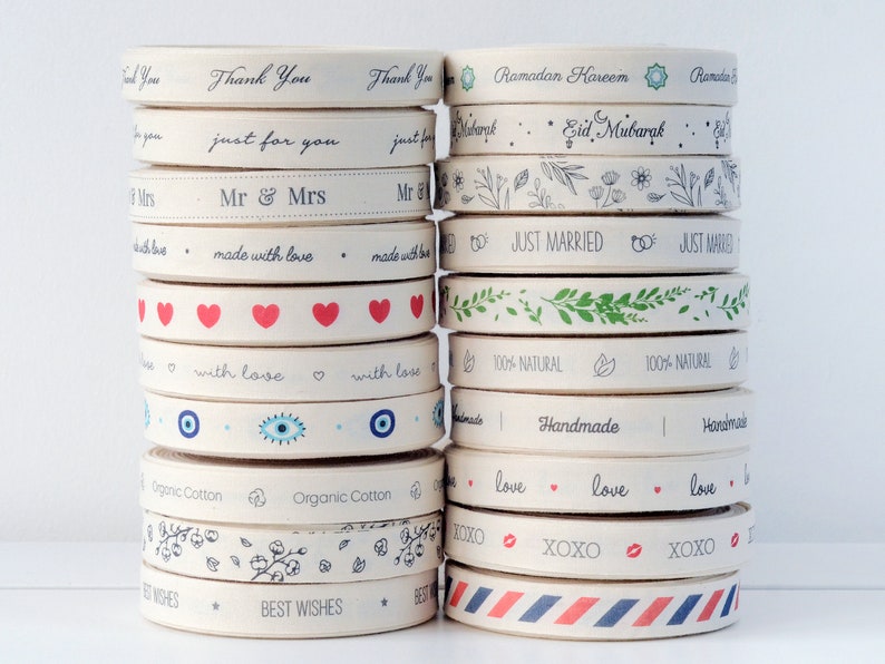 Puede incluir: Un rollo de cinta de algod&oacute;n natural con varios dise&ntilde;os impresos. Los dise&ntilde;os incluyen texto como "Gracias", "Just Married", "Hecho con amor", "Algod&oacute;n org&aacute;nico" y "Mejores deseos". Algunos rollos tienen patrones simples como corazones, hojas y estrellas.