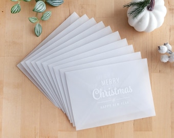 May include: A stack of white translucent envelopes with the words "MERRY Christmas & HAPPY NEW YEAR" printed on the front. The envelopes are arranged on a light brown wooden surface, with decorative elements.