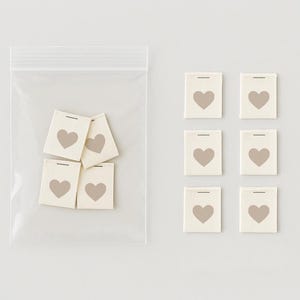 Può includere: Un set di otto etichette in tessuto bianco con un design a forma di cuore. Le etichette sono in un sacchetto di plastica trasparente e su uno sfondo bianco.