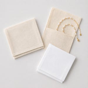 May include: An overhead shot shows a collection of jewelry pouches and cleaning cloths. Two beige fabric pouches are visible, one open with a gold and pearl necklace inside. Two stacks of square cloths, one beige and one white, are also present.