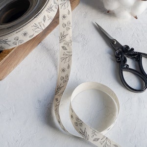 Puede incluir: Un rollo de cinta de tela blanca con un dise&ntilde;o de estampado floral negro. La cinta est&aacute; parcialmente desenrollada y se encuentra sobre una superficie blanca. Tambi&eacute;n hay un par de tijeras decorativas negras sobre la superficie.