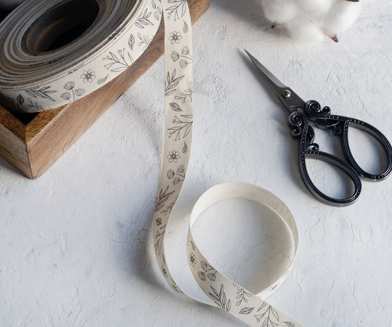 Puede incluir: Un rollo de cinta de tela blanca con un dise&ntilde;o de estampado floral negro. La cinta est&aacute; parcialmente desenrollada y se encuentra sobre una superficie blanca. Tambi&eacute;n hay un par de tijeras decorativas negras sobre la superficie.