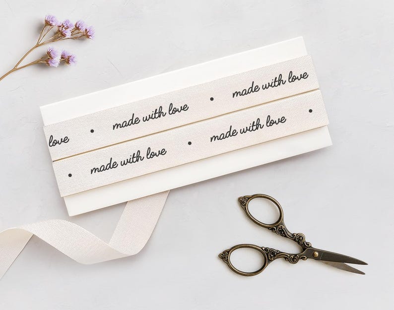 May include: A close-up shot features cream-colored ribbons with the words "made with love" printed in black. The ribbons are wrapped around a stack of white cards. A pair of antique scissors and a sprig of purple flowers are also present.