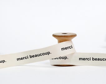 May include: A spool of white ribbon with the text "merci beaucoup." printed on it. The ribbon is wrapped around a wooden spool.