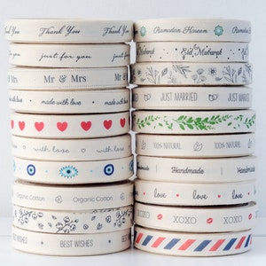 May include: A stack of beige ribbon rolls with various printed designs and text. The ribbons feature phrases like "Thank You," "Just Married," and "Best Wishes," along with heart and floral patterns. The ribbons are made of organic cotton.
