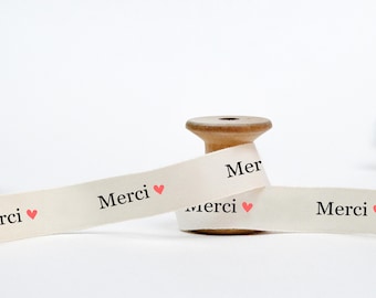 May include: White ribbon with the word "Merci" and a small pink heart printed on it. The ribbon is wrapped around a wooden spool.