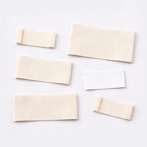 May include: A collection of rectangular labels in different sizes, predominantly in a light beige hue, with one white label. Two of the beige labels feature rolled edges, resembling scrolls. The labels are arranged against a plain white backdrop.