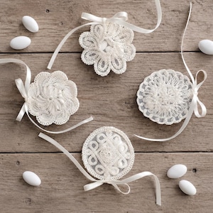 Può includere: Quattro sacchetti in pizzo bianco con intricati motivi floreali e geometrici, ciascuno legato con un nastro di raso bianco abbinato. I contenitori circolari in pizzo contengono piccoli confetti bianchi e alcuni confetti bianchi sono sparsi intorno.