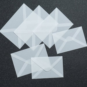 May include: A stack of six white translucent envelopes with a flap on the top. The envelopes are arranged in a staggered pattern.