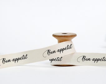 May include: A wooden spool with cream-colored ribbon. The ribbon is printed with the words "Bon appétit" in a cursive font. The ribbon is partially unwound, creating a decorative display against a white background.