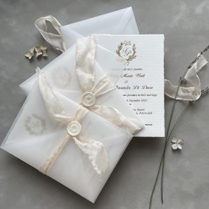 May include: A white wedding invitation with a gold monogram and a white ribbon. The invitation is in a clear envelope with a white wax seal.