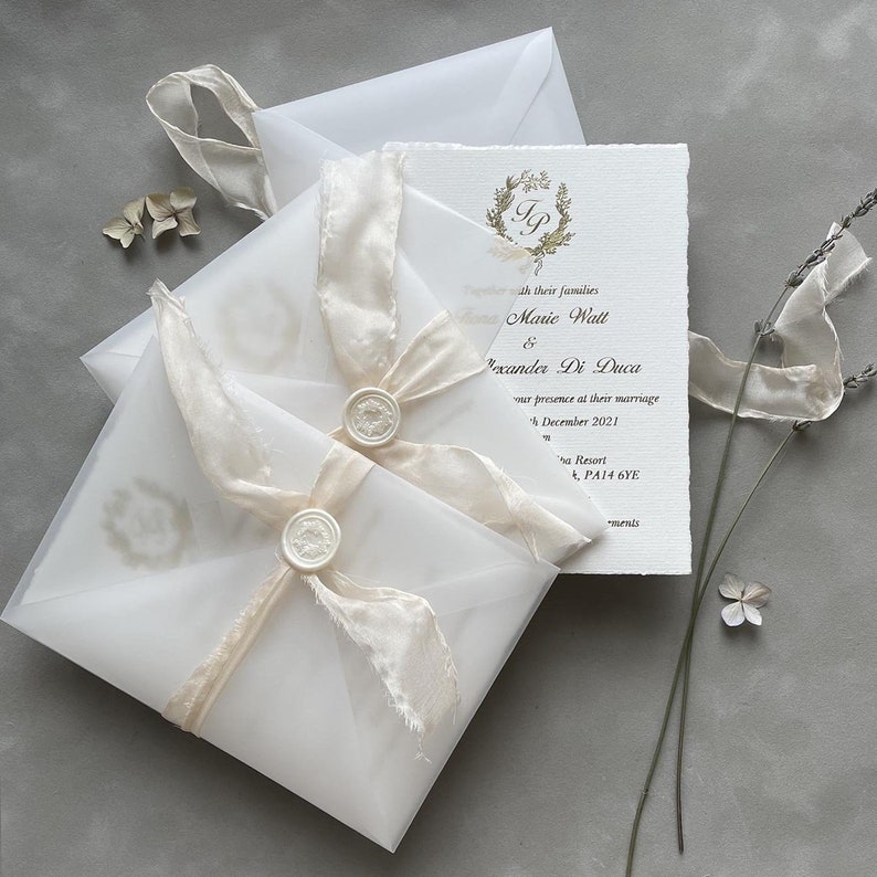 May include: A white wedding invitation with a gold monogram and a white ribbon. The invitation is in a clear envelope with a white wax seal.