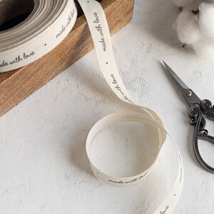 May include: A roll of off-white ribbon with the words "made with love" printed in black. The ribbon is partially unrolled, with a pair of decorative scissors and a wooden box in the background. Cotton buds are also visible.