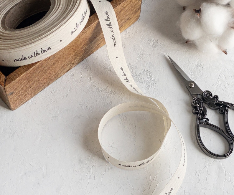 May include: A roll of off-white ribbon with the words "made with love" printed in black. The ribbon is partially unrolled, with a pair of decorative scissors and a wooden box in the background. Cotton buds are also visible.