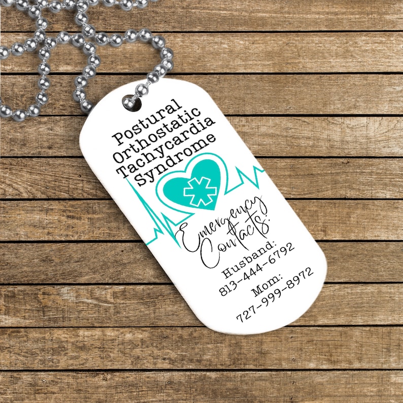 Puede incluir: Collar con placa de alerta m&eacute;dica blanca con un coraz&oacute;n azul y un s&iacute;mbolo m&eacute;dico. La placa dice "S&iacute;ndrome de taquicardia ortost&aacute;tica postural" y "Contactos de emergencia: Esposo: 813-444-6792 Mam&aacute;: 727-999-8972".