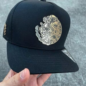 Peut inclure: Casquette de baseball noire ornée d'un écusson doré complexe. L'écusson circulaire présente une figure centrale et des motifs. Un petit écusson doré est visible sur le côté de la casquette.