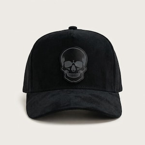 May include: A black baseball cap with a textured, suede-like finish. The front panel features a centered, dark grey graphic patch depicting a detailed human skull. The cap has a structured crown, a curved brim, and a small button at the top center.