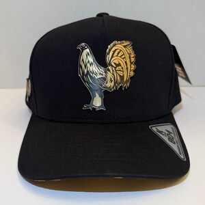 Könnte beinhalten: Schwarze Baseballkappe mit einer detaillierten Illustration eines Hahns in Gold und Weiß. Die Kappe hat einen gebogenen Schirm und eine strukturierte Krone. Das Hahn-Design ist mittig auf der Vorderseite platziert.