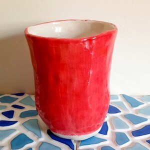 Peut inclure: Un vase en céramique rouge vif avec un bord blanc. Le vase a une surface texturée, légèrement irrégulière et une forme arrondie.