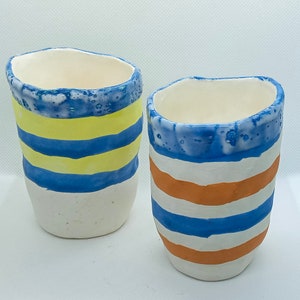 Puede incluir: Dos tazas de cerámica con diseños a rayas. Una taza tiene rayas azules y amarillas, la otra tiene rayas azules y naranjas.