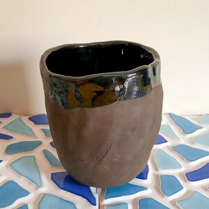 Puede incluir: Una taza de cerámica marrón oscuro con un borde negro esmaltado. La taza tiene un aspecto rústico y hecho a mano.