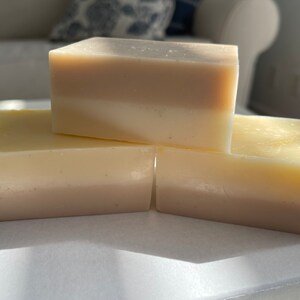 May include: Three bars of handmade soap, each with two layers of different shades of white and beige. The soap bars are stacked on top of each other on a white surface.