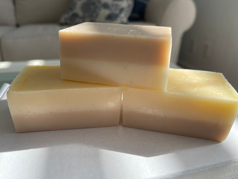 May include: Three bars of handmade soap, each with two layers of different shades of white and beige. The soap bars are stacked on top of each other on a white surface.