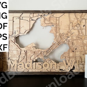 Puede incluir: Un mapa de madera de Madison, Wisconsin, con el nombre de la ciudad recortado y los lagos de la zona también recortados. El mapa está enmarcado en un marco de madera oscura.