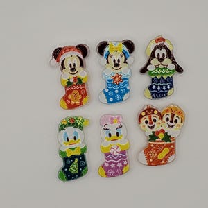 Könnte beinhalten: Ein Set aus sechs bunten Emaille-Anstecknadeln, die Zeichentrickfiguren zeigen, die aus Weihnachtsstrümpfen hervorschauen. Die Motive umfassen Mickey Maus, Minnie Maus, Goofy, Donald Duck, Daisy Duck sowie Chip und Chap mit festlichen Mustern.