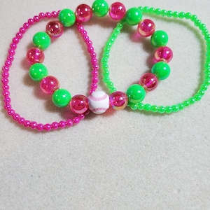 May include: Two elastic bracelets featuring a mix of neon green and translucent pink beads. One bracelet includes a small, round white bead with red stitching resembling a baseball. The other bracelet consists of smaller neon green beads.