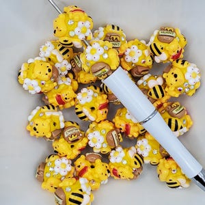 May include: A white pen featuring a stack of yellow bear-shaped beads with white flowers and small rhinestones. Each bead depicts a yellow bear with a brown pot labeled HUNNY and black and yellow striped bee-like elements with white wings.