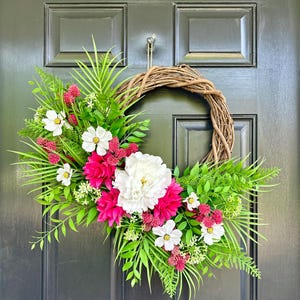 May include: A decorative wreath featuring a woven grapevine base adorned with vibrant faux flowers and greenery. The arrangement includes large white and fuchsia-colored blooms, white daisies, and lush green foliage, creating a welcoming spring or summer aesthetic.