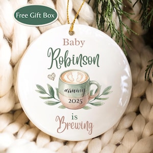 May include: A white ceramic Christmas ornament with gold string. The ornament features the text "Baby Robinson arriving 2025 is Brewing" with a coffee cup design and greenery. A small green box in the upper left corner says "Free Gift Box".