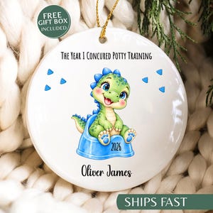 May include: A round white ceramic ornament featuring a green cartoon dinosaur sitting on a blue potty. The text reads The Year I Concured Potty Training, 2026, and Oliver James. Small blue poop icons surround the dinosaur, and a gold string is attached.
